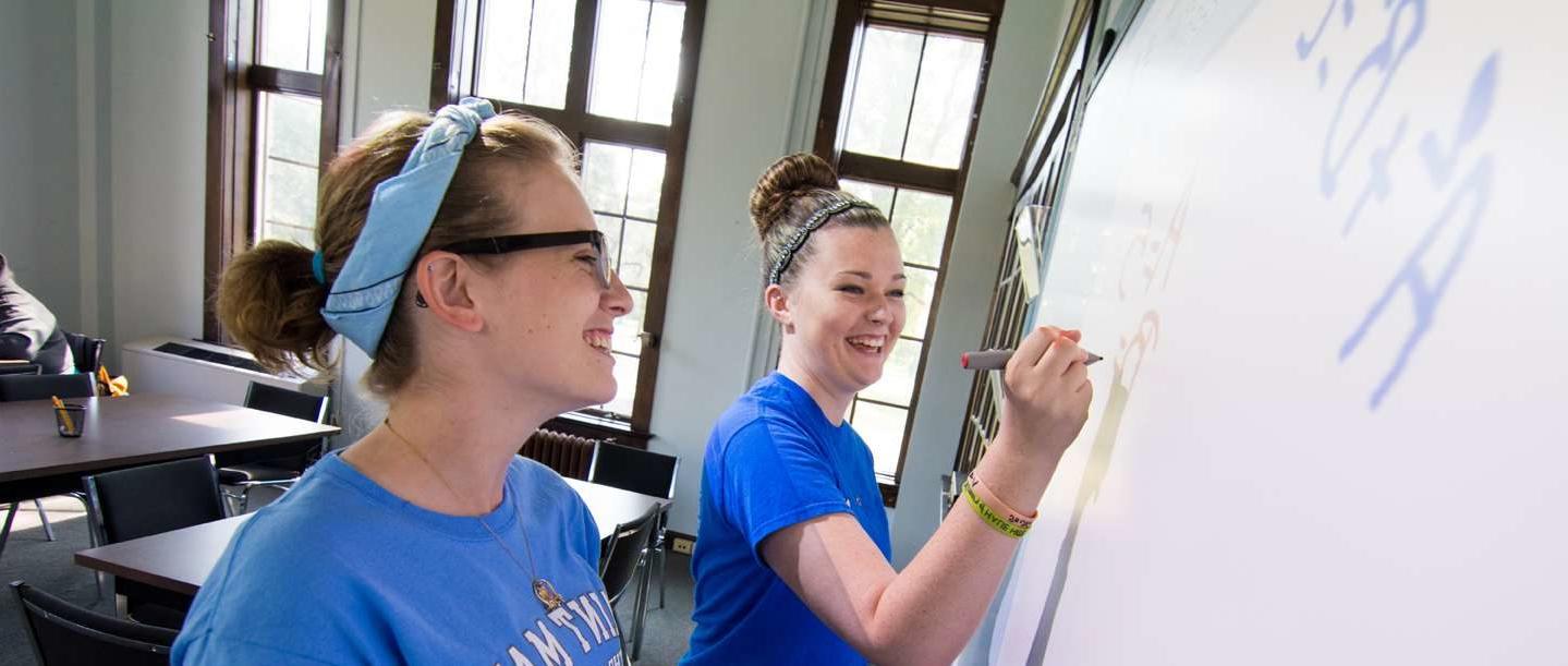 Two female students standing at a dry erase board working together on homework.